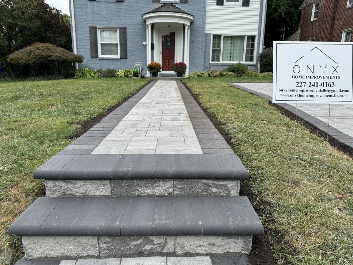 Wide light paver walkway with a dark contrasting border running from capped entry steps to the front door of a blue colonial