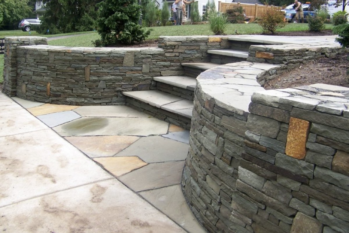 Curved stacked stone retaining wall with capped steps rising between two wall runs to a flagstone landing in a sloped front yard