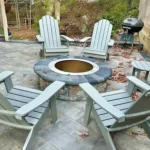 Close up of patio furniture surrounding a natural stone fireplace. Tall trees blanket the background.