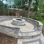 Natural stone fireplace encircled by a circular retaining wall. We see a wooden fence and trees in the background.
