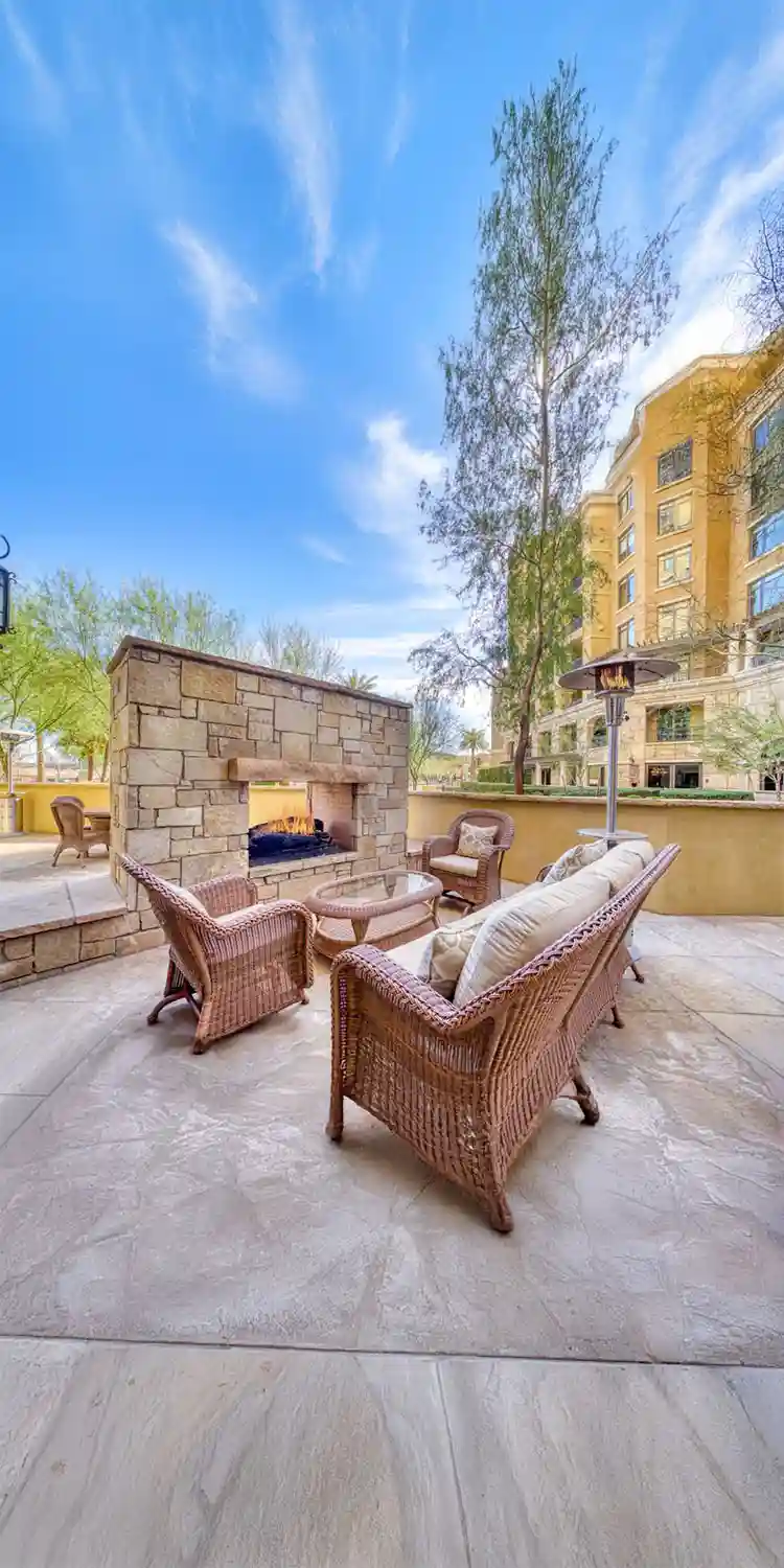 Outdoor stone fireplace with patio furniture in front of it. An apartment building is in the background, semi covered by trees.