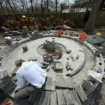 Patio construction underway. A pile of natural stones are on the ground to the side of a male contractor.