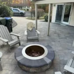 Residential backyard patio made out of natural stone. A stone veneer fireplace sits in the middle of the patio, surrounded by chairs.