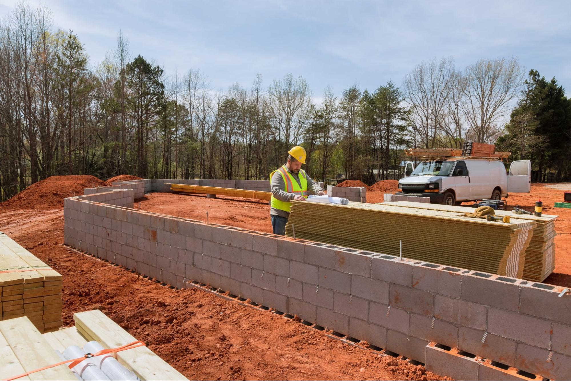 Construction site supervisor wearing a hard hat and safety vest inspects the concrete block foundations.