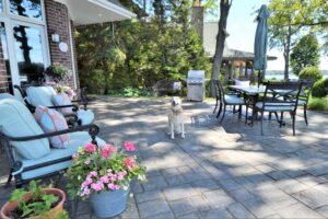 A white dog sits in the middle of a cozy backyard patio with a custom paver design opening into a garden area.