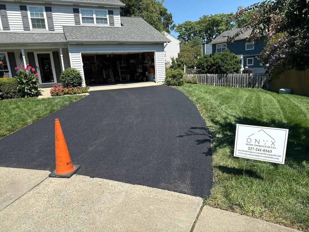 Asphalt driveway paving in progress with orange safety cone at entrance.