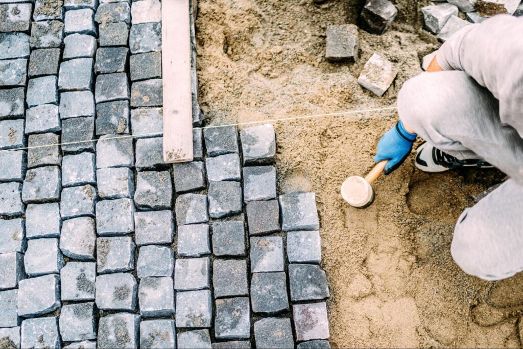 A paving company worker installs pavers on sand, using a rubber mallet.