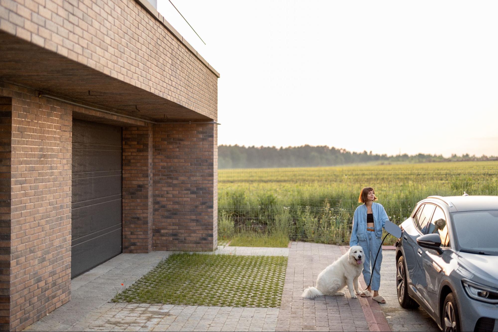 A woman and her dog stand on their driveway while plugging in her electric car.