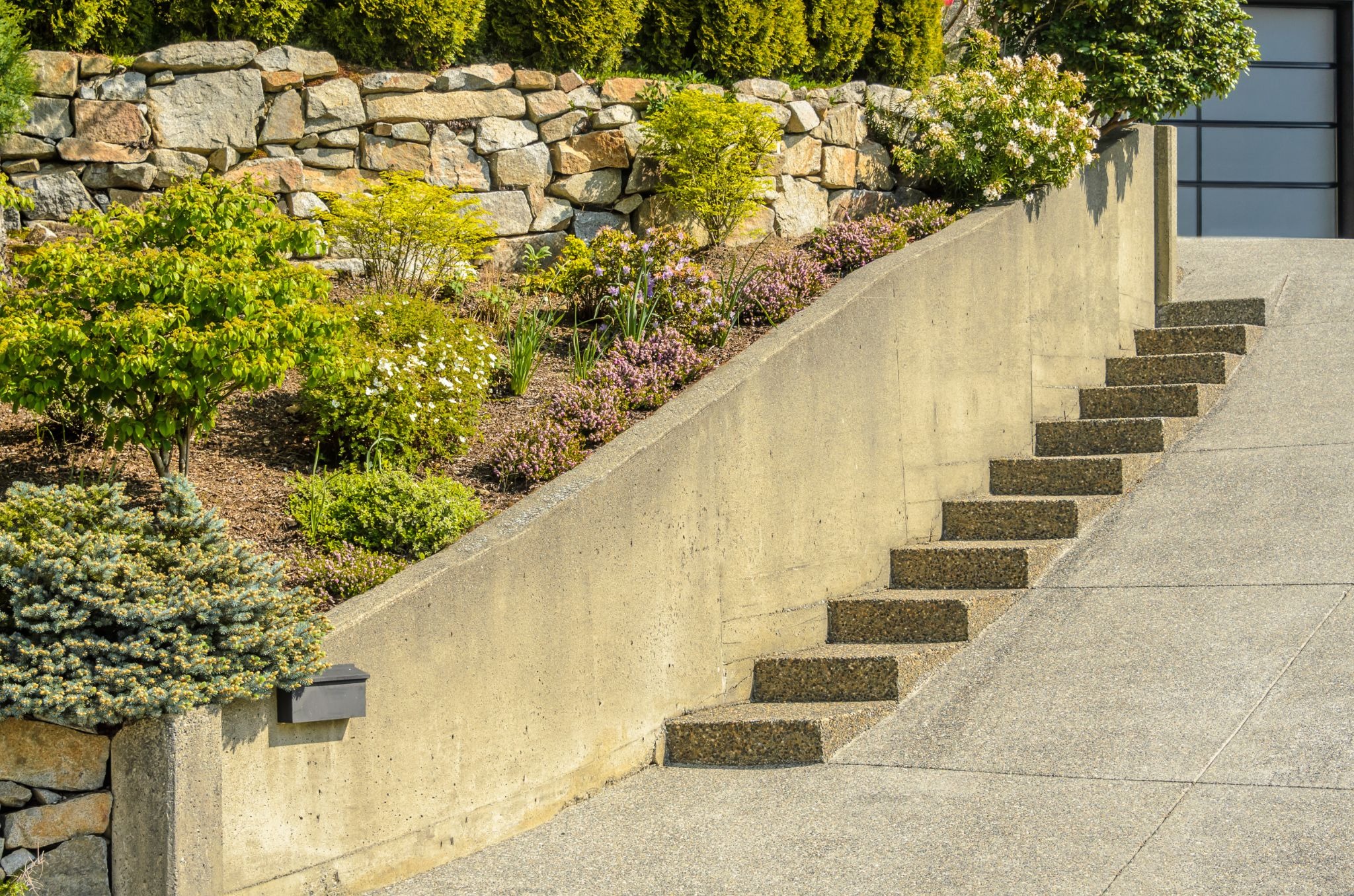 Cement retaining wall with plants and stone steps nearby.